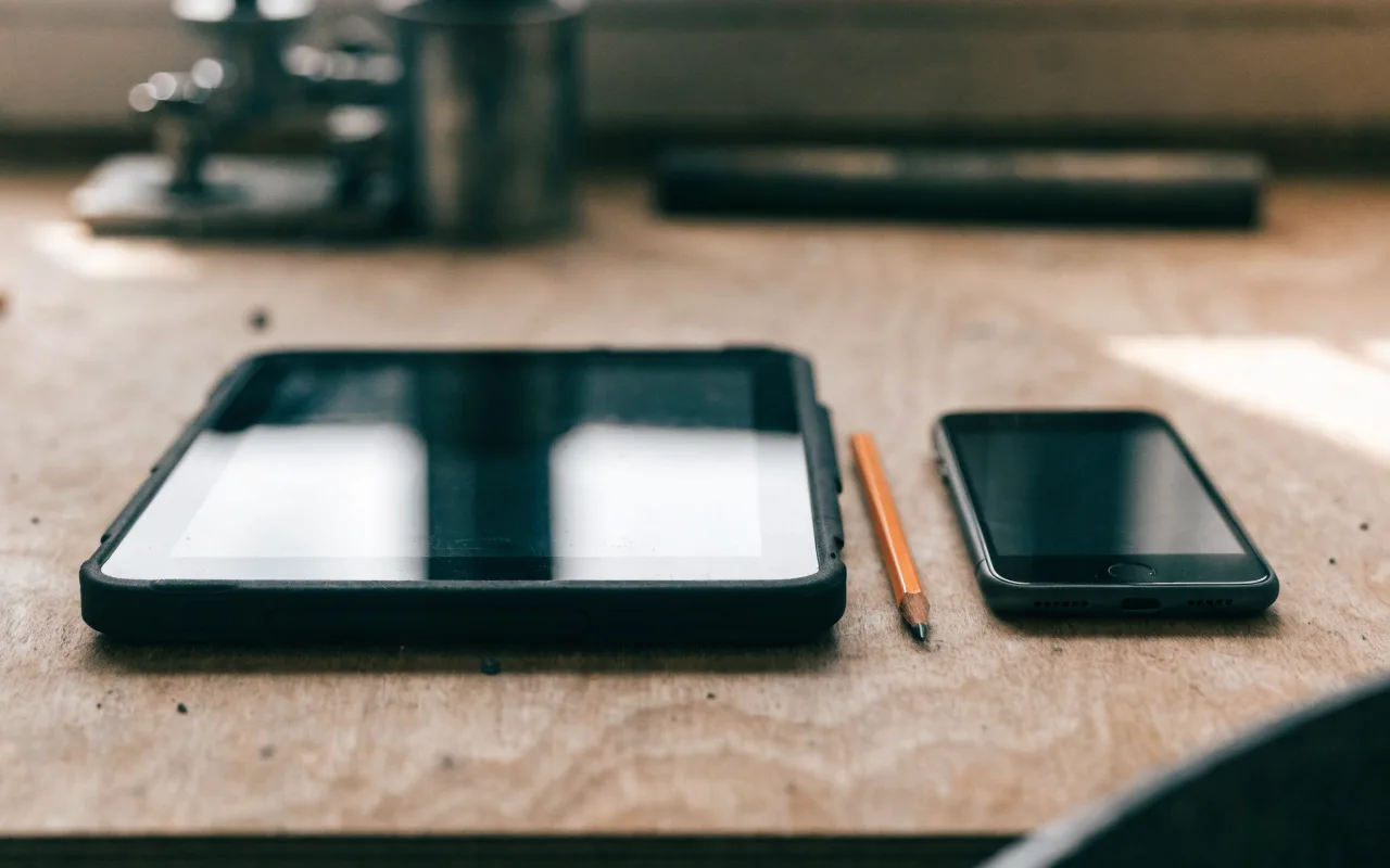 A tradie's iPad and an Android phone side by side on a plywood workbench, an orange pencil between them.