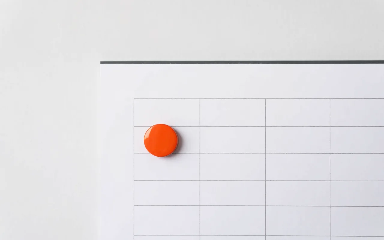A blank white wall planner grid with a single orange magnet pinned on one day.