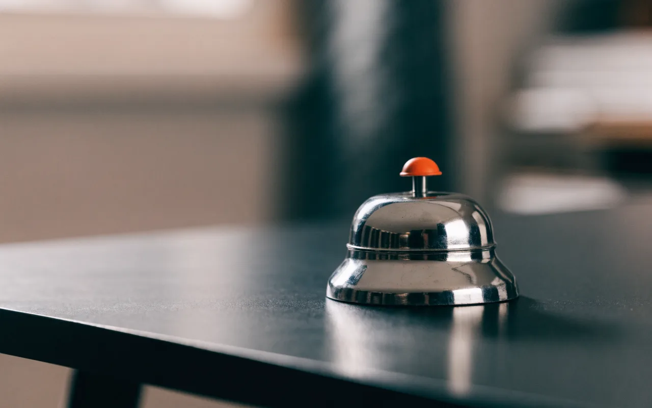 A chrome office desk bell on a dark desk, its press button coloured orange.