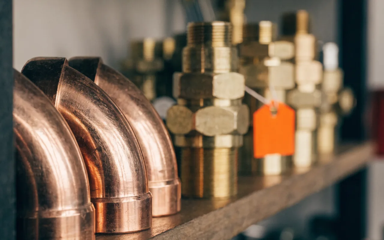 Copper pipe elbows and brass fittings on a plumber's shelf, one fitting wearing an orange price tag.
