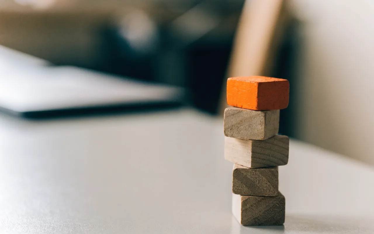 A stack of plain wooden building blocks on a grey desk, the top block painted orange.